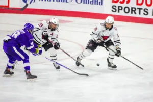 Omaha Mavericks forward Jack Randl (28) with  forward Ty Mueller (19) keeps the puck away from Niagara Purple Eagles forward Tyler Wallace (18) in the third period during a college hockey match on Friday, October 13, 2023, in Omaha, Nebraska. Photo by John S. Peterson.